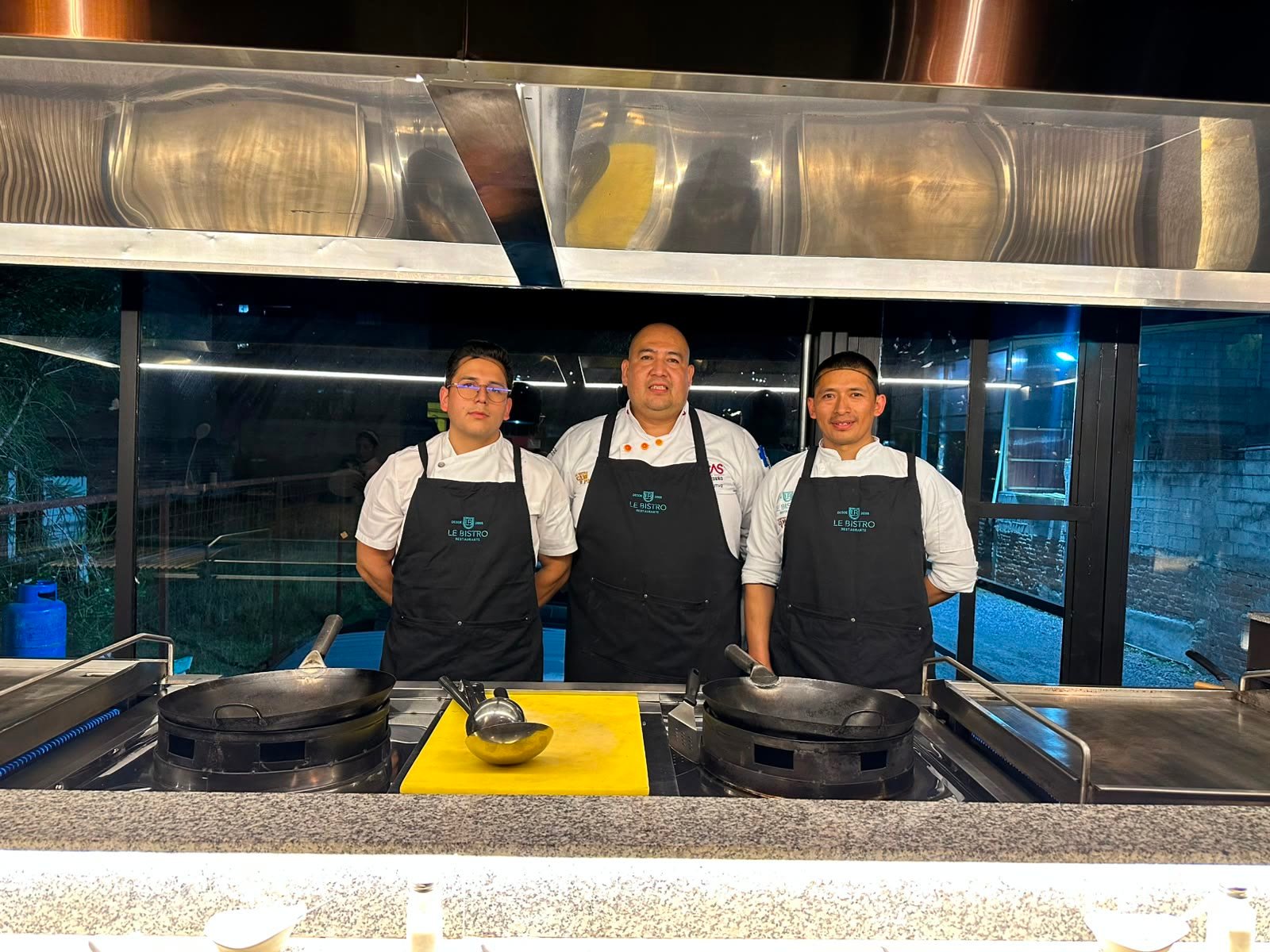 Los tres chefs de Le Bistro de pie detrás de la barra, con delantales negros y los woks listos